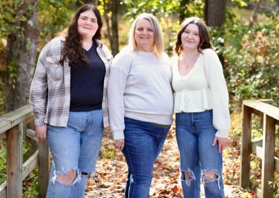 Three people stand together on a wooden bridge in a wooded area with fall foliage.