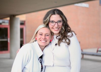 Two smiling people wearing white tops pose closely together outdoors in front of a brick building.