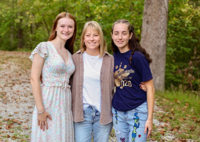 Three people pose together outdoors in a wooded setting, with one wearing a blue t-shirt that reads "Class of 28."