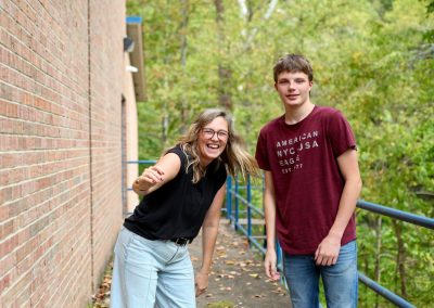A smiling adult and a young person standing on an outdoor walkway next to a brick building, with the person on the right wearing a shirt that says "American NYC USA."